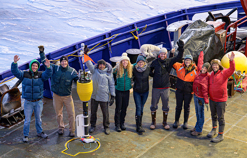Nine people standing on the deck of the Sikuliaq and waving at the camera
