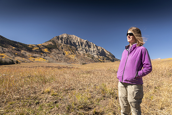 Professor Jessica Lundquist stands in an open field with mountains and fall foliage in the background, wearing sunglasses, a purple jacket, and khaki pants.