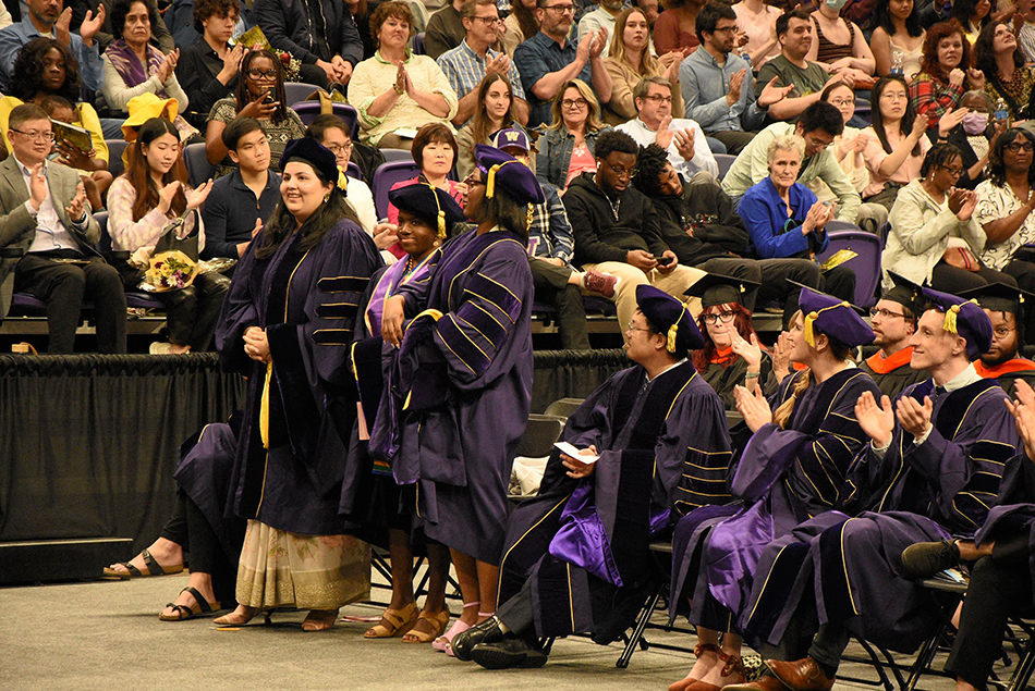 Group of doctoral candidates standing and sitting during the ceremony, applauded by the audience.