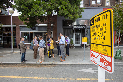 A group of people stands on a sidewalk in an urban area, near a sign indicating parking regulations