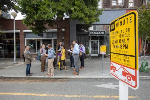 A group of people stands on a sidewalk in an urban area, near a sign indicating parking regulations