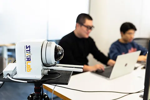 A Mobile Unit for Sensing Traffic (MUST) sensor sitting in the foreground and two researchers working on laptops in the background