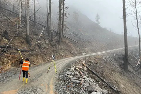 A person in an orange vest and hard hat stands on a dirt road in a forested area with burnt trees, holding surveying equipment. Fog covers the background.