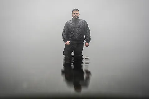 A man stands in calm, mid-thigh-deep water with a foggy background.