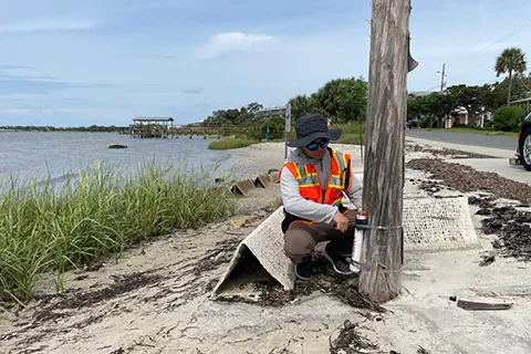 A researcher wearing a high-visibility vest takes measurements from a beach on a sunny day.