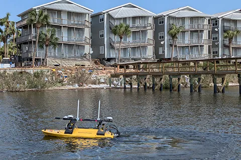 The RAPID team’s Z-boat surveys coastal waters in Florida to map underwater changes caused by Hurricane Helene. In the background, apartments show visible damage from the storm.